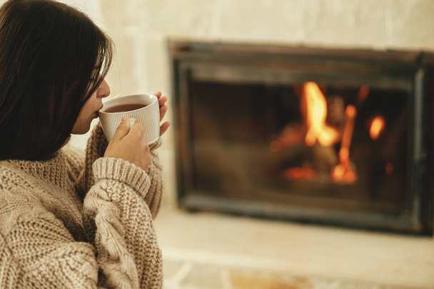 Woman in cozy sweater drinking cup of warm tea at fireplace in rustic room. Heating house in winter with wood burning stove. Young stylish female relaxing at fireplace in farmhouse - Photo, Image