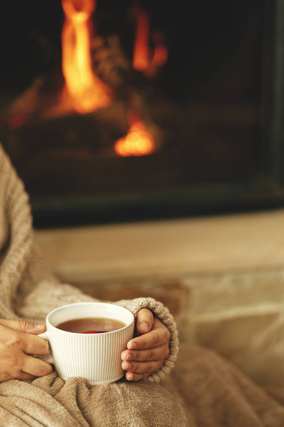Hands in cozy sweater holding cup of warm tea on background of burning fireplace close up, autumn hygge. Relaxing and warming up at rustic fireplace. Heating house with wood burning stove - Photo, Image