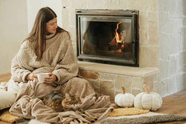 Woman with cup of tea and cute cat sitting together on cozy blanket at fireplace. Adorable tabby kitty relaxing together with owner at burning fireplace in rustic farmhouse - Photo, Image