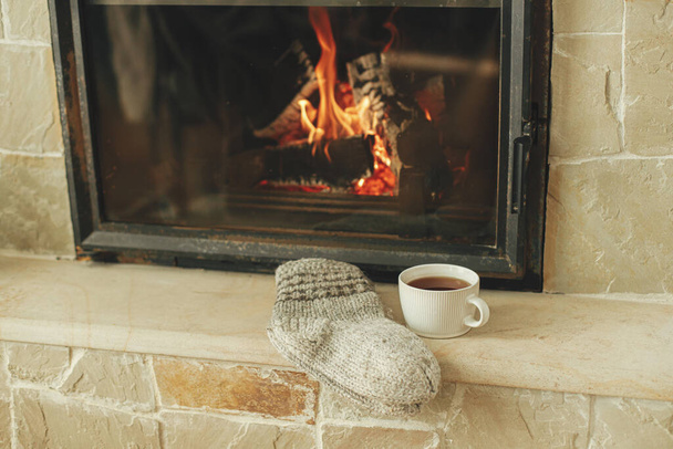 Stylish cup of warm tea and cozy socks on background of burning fireplace close up, autumn winter hygge. Heating house with wood burning stove. Relaxing and warming up at rustic fireplace - Photo, Image