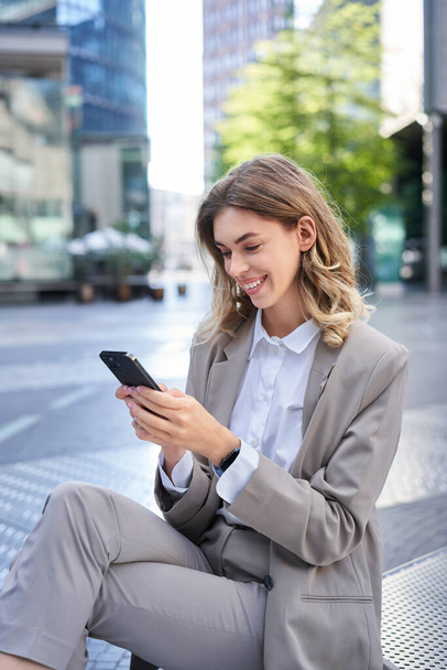 Verticale opname van glimlachende zakenvrouw op zoek naar smartphone app, verzenden van bericht op mobiele telefoon, oder lunch tijdens het zitten buiten kantoorgebouw. - Foto, afbeelding