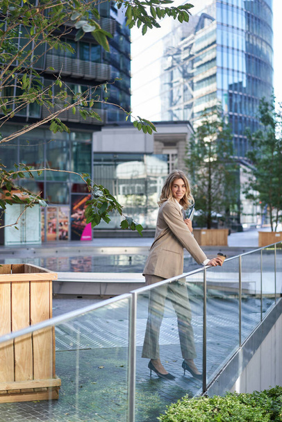 Vertikale Aufnahme einer Geschäftsfrau im beigen Anzug, die draußen auf der Straße steht und vor der Arbeit Kaffee zum Mitnehmen trinkt. - Foto, Bild