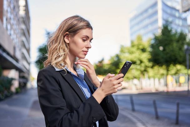 Profile portrait of young saleswoman, corporate lady in suit, using mobile phone, reading on smartphone, standing on street outside office. - Photo, Image