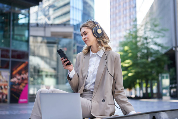 Mujer de negocios sonriente en traje, sentada en una reunión en línea desde el portátil, usando auriculares, mirando su teléfono móvil con cara feliz. - Foto, Imagen