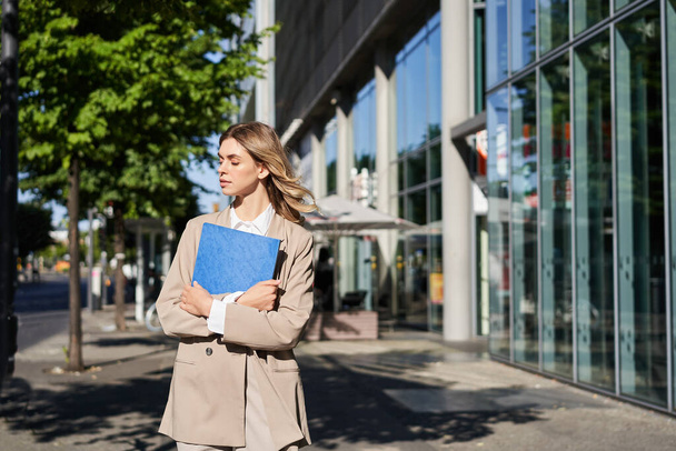 Trabajo y gente corporativa. Mujer de negocios confiada sosteniendo una carpeta azul con documentos de negocios, caminando por la calle. - Foto, imagen