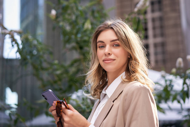 Close up portret uśmiechniętej bizneswoman, młody stażysta biurowy w garniturze, trzymając smartfona i patrząc szczęśliwy przed kamerą, stojąc na ulicy. - Zdjęcie, obraz