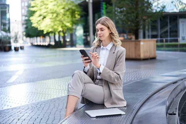 Zakelijke vrouw zit in het centrum van de stad en werkt op mobiele telefoon, ze heeft haar laptop en draagt werkende zakelijke outfit. - Foto, afbeelding