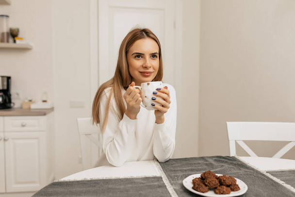 Cute adorable woman with blond hair wearing white shirt is sitting at kitchen Wirth happy smile. Caucasian woman resting at home with coffee and cookies  - Photo, image