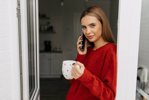 Schattig charmant meisje met blond haar draagt rode trui is praten aan de telefoon en holding cup met koffie inn de ochtend in de keuken  - Foto, afbeelding