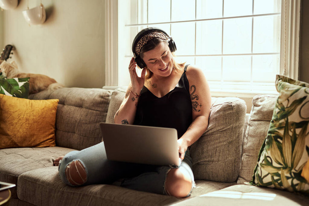 Έτσι ακούγεται η ευτυχία. μια νεαρή γυναίκα που χρησιμοποιεί ένα φορητό υπολογιστή και ακουστικά στον καναπέ στο σπίτι - Φωτογραφία, εικόνα