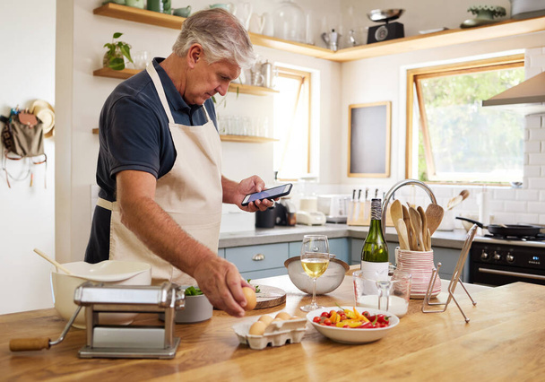 Senior man, cooking and phone for recipes, research or ingredients for the perfect meal in the kitchen at home. Elderly male cook or baker with food browsing online recipe or streaming on smartphone. - Photo, image