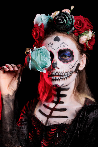 Studyjne ujęcie młodej kobiety z makijażem czaszki, wyglądającej olśniewająco z kwiatową koroną i body artem. Świętowanie dnia martwych meksykańskich świąt z różami i przerażającą tradycją festiwalową. - Zdjęcie, obraz