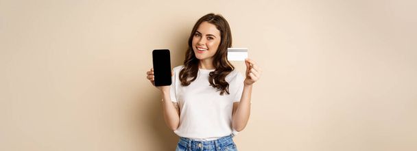 Online shopping and people concept. Young beautiful woman looking happy, showing credit card discount and mobile phone screen, standing over beige background. - Fotó, kép