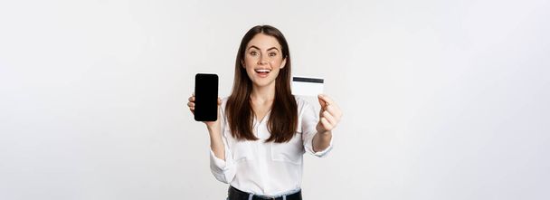 Portrait of female model showing credit card with smartphone screen, recommending application, standing over white background. - Foto, immagini