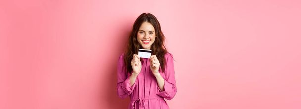 Красивая молодая женщина с пластиковой кредиткой и улыбкой, покупки в магазинах, стоя на розовом фоне. - Фото, изображение