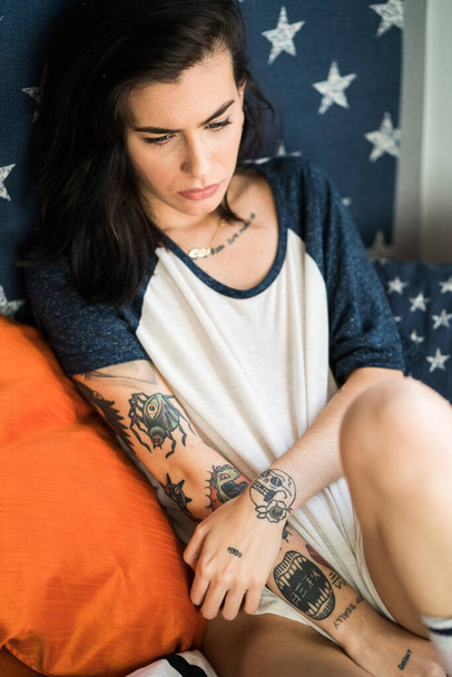 Young woman with tattoos sitting on a sofa with a united states flag on wall relaxing - Photo, image