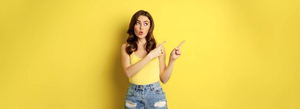 スタイリッシュな夏の女の子の肖像画、ブルネットの女性が道を示す、指を左に指して、広告、黄色の背景の上に立っている。スペースのコピー - 写真・画像