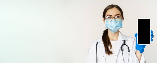 Online medische hulp concept. Vrouwelijke arts in bril en gezichtsmasker, toont mobiele telefoon scherm, app-interface of website voor patiënten, staande over witte achtergrond. - Foto, afbeelding