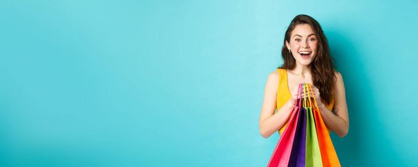 Νεαρή όμορφη γυναίκα κρατώντας τσάντες για ψώνια, χαμογελώντας ενθουσιασμένοι με την κάμερα, αγοράζοντας με εκπτώσεις, στέκεται πάνω από το μπλε φόντο. - Φωτογραφία, εικόνα