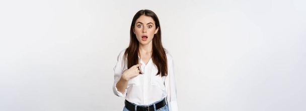 Portrait of young woman pointing at herself with disbelief, surprised face expression, being named or chosen, standing over white background. - 写真・画像