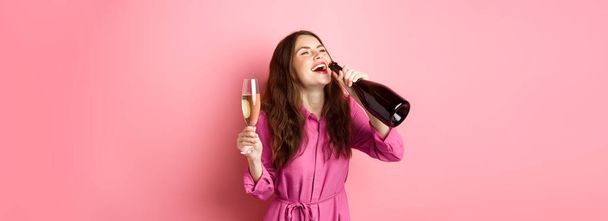 Ünnepség és ünnepek koncepció. Boldog gondtalan nő iszik pezsgőt üveg elégedett mosollyal, pihentető partin, kezében üveg, álló felett rózsaszín háttér. - Fotó, kép