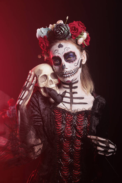Piękna kobieta w tradycyjnym makijażu, aby świętować meksykańskie święto, trzymając czaszkę i czarne róże w studio. Wygląda jak Santa Muerte lub bogini śmierci na świętych dios de los muertos. - Zdjęcie, obraz