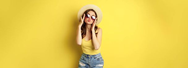 Happy sexy young woman on vacation, wearing straw hat, sunglasses, laughing and smiling, standing over yellow background. - Photo, Image