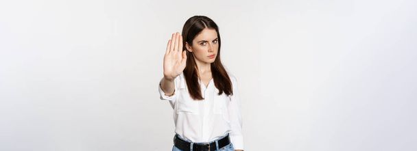 Серьезный женщина показывает стоп-знак, не согласен, отвергая smth плохо, стоя на белом фоне. - Фото, изображение
