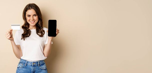 Online shopping and people concept. Young beautiful woman looking happy, showing credit card discount and mobile phone screen, standing over beige background. - 写真・画像