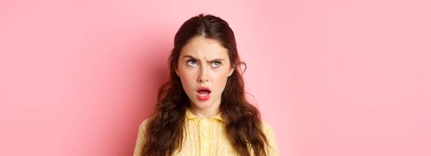 Young brunette woman with confused face, trying figure something out, frowning and looking up while thinking, standing pensive against pink background. - Photo, Image