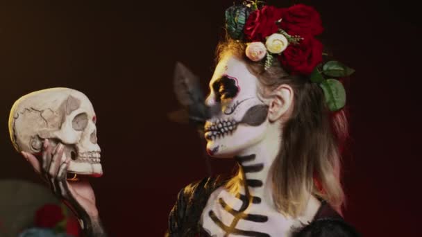 Flirtující mrtvá dáma se chová děsivě s lebkou v ruce, drží černé růže a nosí děsivý make-up. Vypadá to jako Santa Muerte s tělesným uměním na svaté mexické oslavě, být okouzlující. Ruční snímek. - Záběry, video