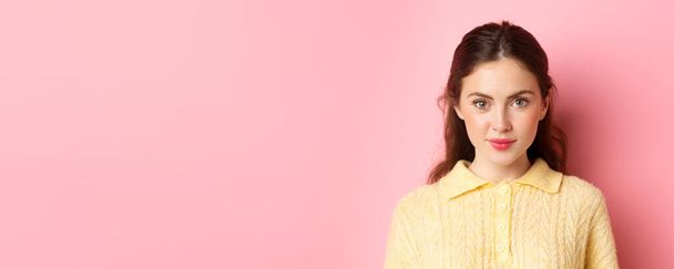 Κοντινό πλάνο του νεαρού κορίτσι αίγλη με χαριτωμένο μακιγιάζ, κοιτάζοντας αποφασισμένη κάμερα, χαμογελώντας, στέκεται πάνω σε ροζ φόντο. - Φωτογραφία, εικόνα