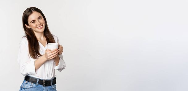 Corporate woman standing with coffee tea mug and smiling, drinking from cup, standing happy against white background. - Photo, image