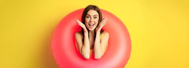 Happy beautiful young woman relaxing on summer vacation, wearing pink swim ring for beach holiday, standing against yellow background. - Фото, изображение