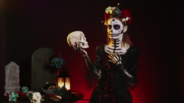 Modelo de terror espeluznante posando con cráneo en estudio, actuando espeluznante en halloween mexicano dios de los muertos. Mujer coqueta que luce como la cavalera catrina para celebrar la cultura tradicional. - Imágenes, Vídeo