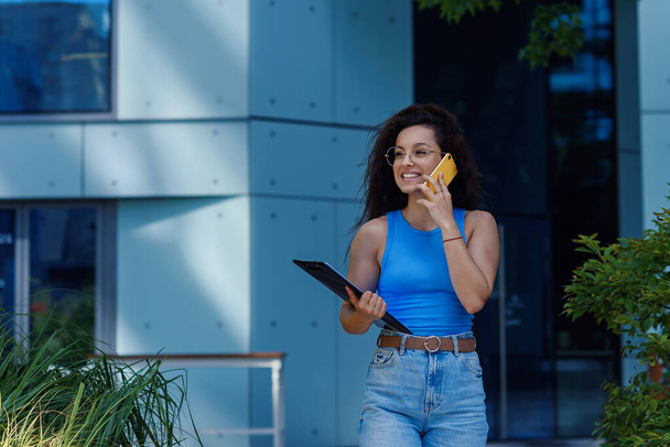 Successful beautiful smiling business woman with document speaking by phone near modern office building. Happy office worker concept. - Фото, изображение