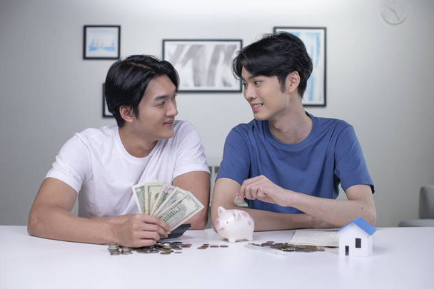 Νεαρό ευτυχισμένο ζευγάρι Ασιατών γκέι που μαζεύει λεφτά για να αγοράσει σπίτι. ΛΟΑΤ άνδρες ζευγάρι εξοικονόμηση χρημάτων για την οικογένεια και το σπίτι. - Φωτογραφία, εικόνα