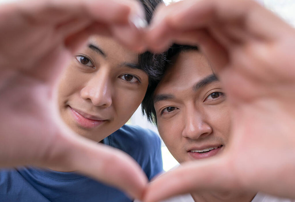 Nuori aasialainen homo pari romanttisella tuulella tehden sydämen muotoinen käsi ele. LGBT miespari osoittaa rakkautta ja kiintymystä pelatessaan kameran kanssa kotiympäristössä. - Valokuva, kuva