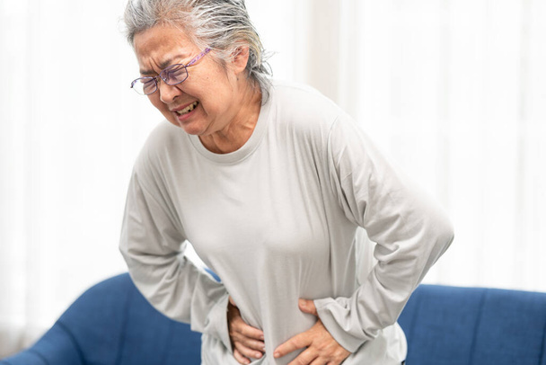 Vanhempi nainen, joka kärsii vatsakivuista kotona. Tarttuminen ja puristamalla vatsa käsin, tunne väsynyt, seisoo olohuoneessa. Ilmaiseva kipu kasvoilla - Valokuva, kuva