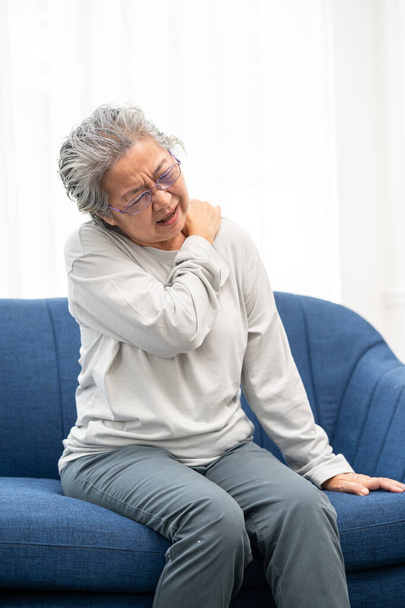 Idősebb nő otthon ül egy kanapén, vállfájdalommal, fájdalommal és rosszullét kifejezésével. Egészségügyi probléma fogalma. - Fotó, kép