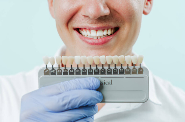 Boca sonriente de hombre con dientes blancos naturales en fondo azul claro en clínica dental. Las manos sosteniendo la paleta de colores de los dientes cara a cara. Sonríe sano concepto de dientes - Foto, imagen