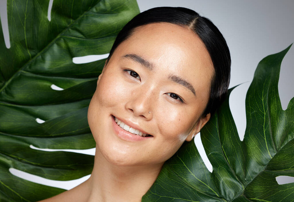 Face, beauty and asian woman with monstera leaf on gray studio background. A beautiful Japanese woman with healthy, glowing and flawless skin, skincare routine and makeup with organic plant cosmetics. - Foto, afbeelding