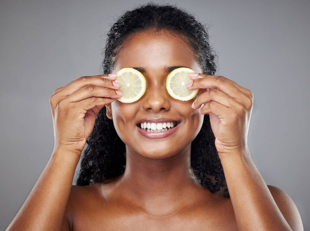 Skincare, portrait and woman with lemon on her eyes for health, beauty and wellness standing in a studio. Happy, smile and face of girl model from Brazil with citrus fruit isolated by gray background. - Fotoğraf, Görsel