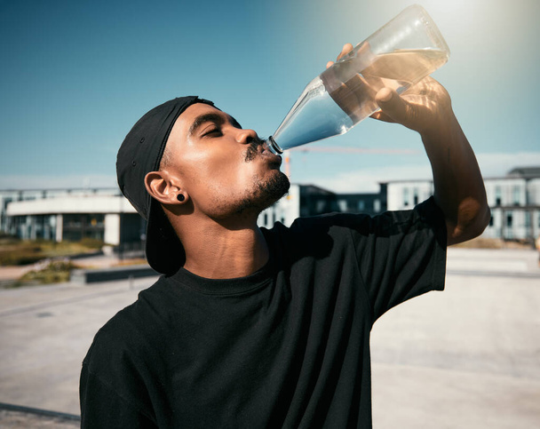 City heat, summer and black man drinking water on concrete road, thirsty gen z outdoor activity. Sun, fun and urban young man with healthy street culture lifestyle and clean water in bottle to drink - Photo, Image