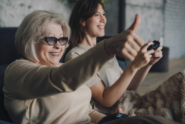 Elderly pensioner playing games on video console showing the like sign, rejoicing that she won. Game win. Modern game joystick. Happy family. - Photo, Image