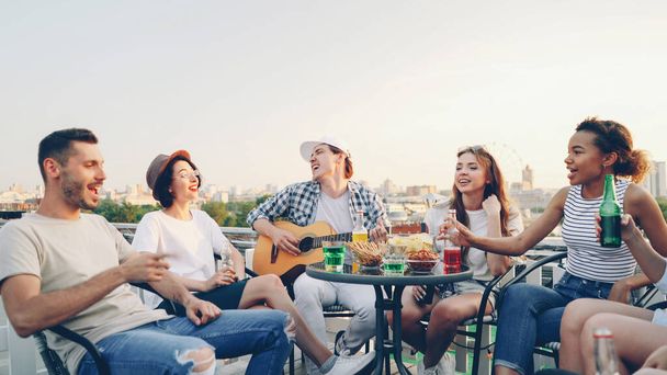 Creative guy in casual clothes is playing the guitar and his friends multi-ethnic group are singing holding bottles with beer and soft drinks sitting in circle at table on roof. - Photo, Image