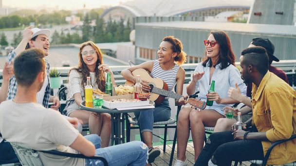 Pretty African American girl is playing the guitar while her happy friends multiethnic group are singing and moving hands enjoying party on rooftop. Table with food and drinks is visible. - Foto, imagen