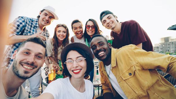 Point of view shot of pretty young lady in trendy hat holding camera and taking selfie with friends enjoying rooftop party with soft drinks, people are looking at camera and laughing. - Foto, imagen
