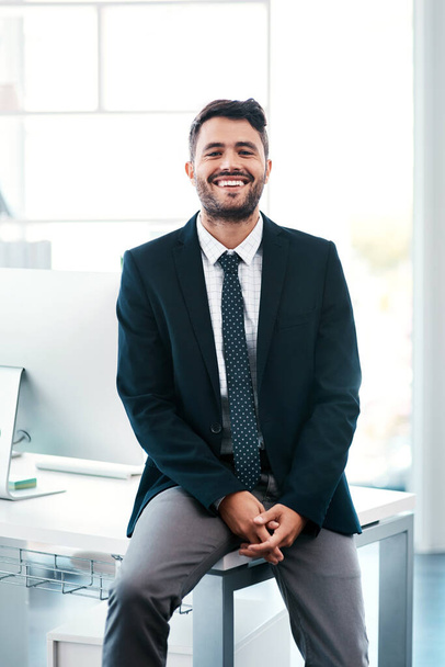 Hard work got me here. Cropped portrait of a handsome young businessman smiling while sitting on his desk in a modern office - Photo, Image