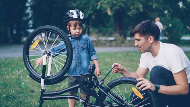 Curious child wearing helmet is spinning bicycle wheel and pedals while his careful father is talking to him on lawn in park on summer day. Family, leisure and active lifestyle concept. - Fotó, kép